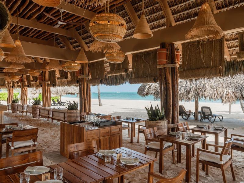 Beachside restaurant with wooden tables, chairs, woven lamps, and ocean views under a thatched roof.