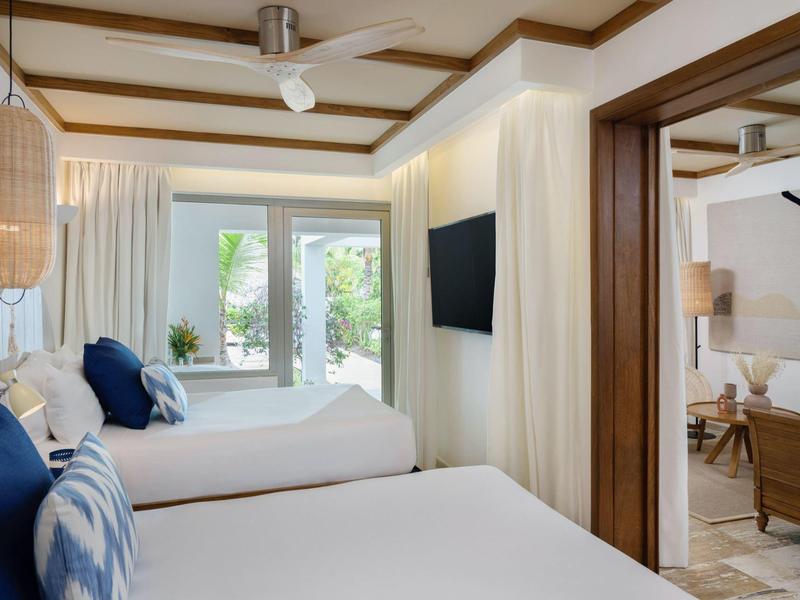 Modern hotel room with two beds, blue pillows, ceiling fan, and view into lounge area.