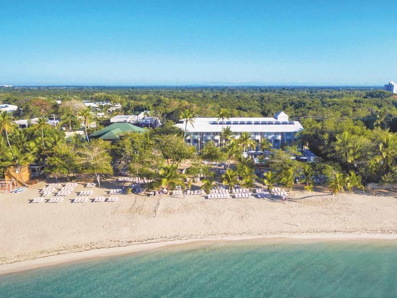 Strand mit hellem Sand, ruhigem blaugrünem Wasser und bewaldetem Ufer unter klarem Himmel.