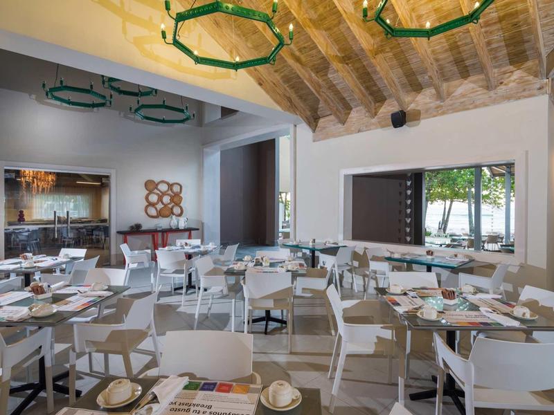 Moderner Gastraum mit weißen Stühlen, Holztischplatten, grünen Deckenlampen und großen Fenstern.