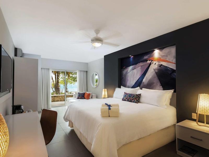 Modernes Hotelzimmer mit großem Doppelbett, Sitzecke und Blick durch Fenster auf Garten