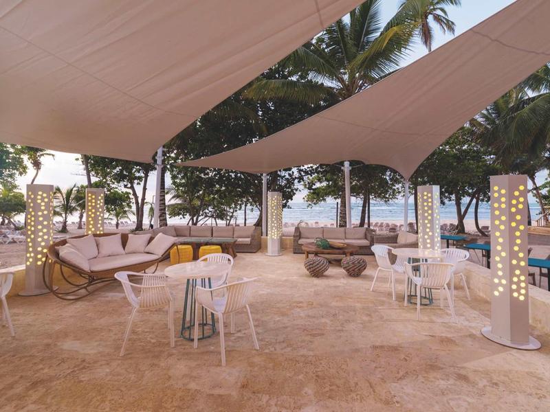 Offener Strandbereich mit weißen Segeltüchern, Stühlen, Tischen und Palmen im Hintergrund.