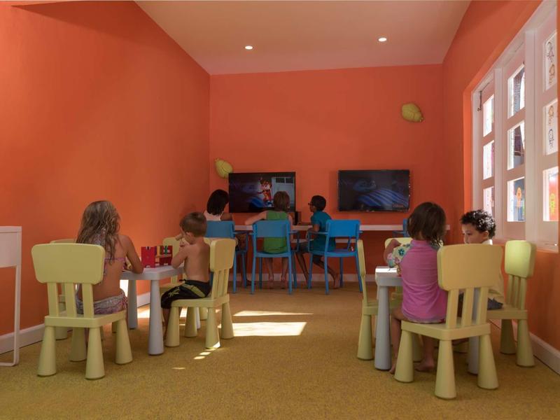 Kinder sitzen an kleinen Tischen in einem hellen, orangefarbenen Raum mit Fenstern und Fernsehern.