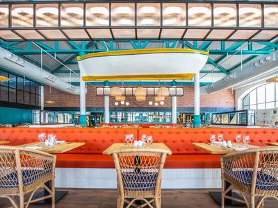 Modernes Restaurant mit orangen Sitzbänken, weißen Holztischen und Flechtstühlen bei großem Fenster.