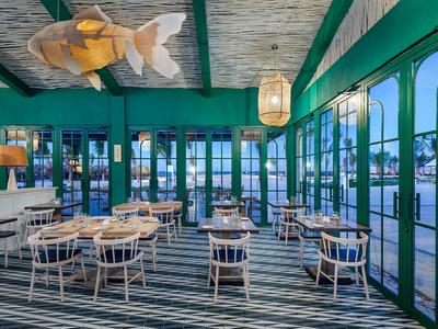 Offenes Restaurant mit grünem Stahlrahmen, Holztischen, weißen Stühlen und Holzlamellen-Decke.