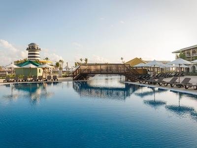 Large outdoor pool with sun loungers and bridge under blue sky at a hotel resort.