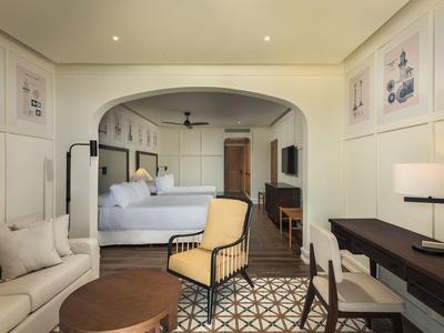 Modernes Hotelzimmer mit offenem Grundriss, hellem Sofa, Sessel, Schreibtisch und einem großen Bett.