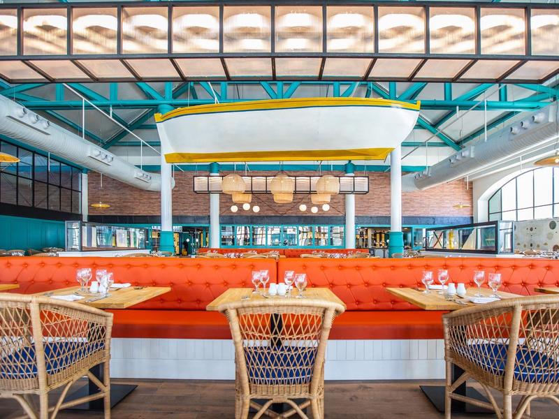 Modernes Restaurant mit orangen Sitzbänken, weißen Holztischen und Flechtstühlen bei großem Fenster.