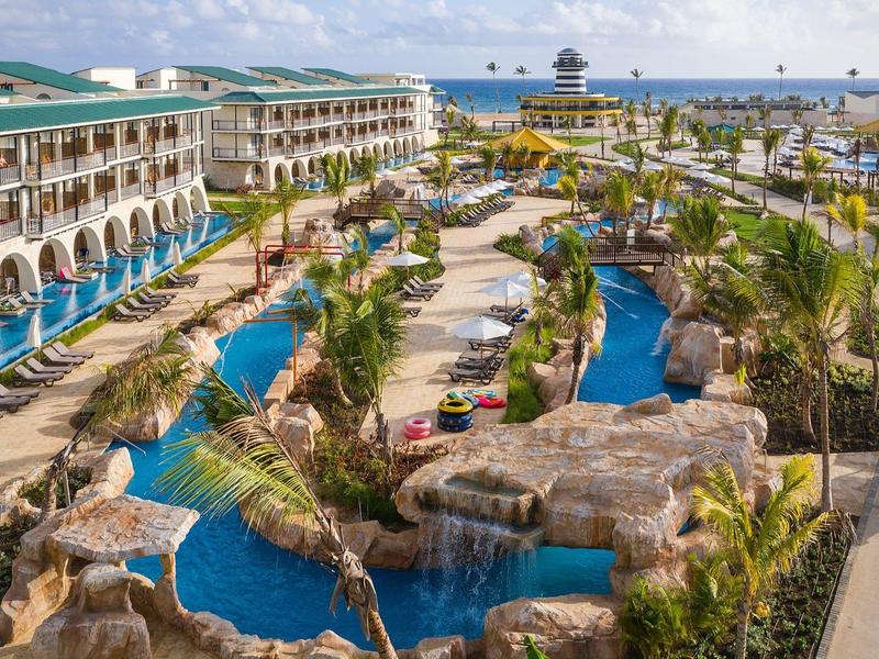 Großes Resort mit mehreren Gebäuden, Palmen, Pools, künstlichen Wasserläufen und Blick aufs Meer.