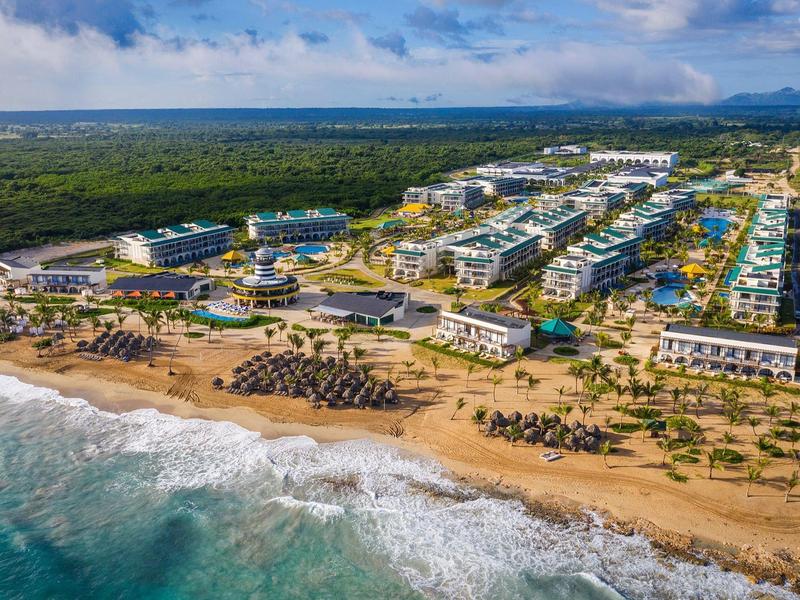 Luftaufnahme eines Küstenresorts mit Sandstrand, vielen Gebäuden und grünem Wald im Hintergrund.