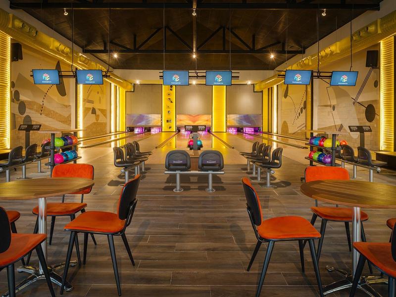 Innenansicht einer modernen Bowlingbahn mit Tischen und Stühlen im Vordergrund.