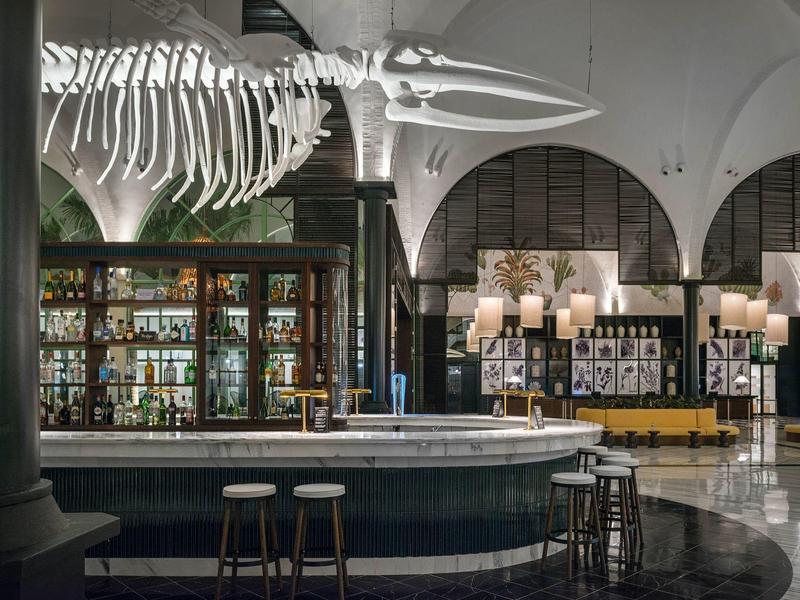 Eleganter Hotelbarbereich mit rundem Tresen, Barhockern und großem Skelett über der Bar