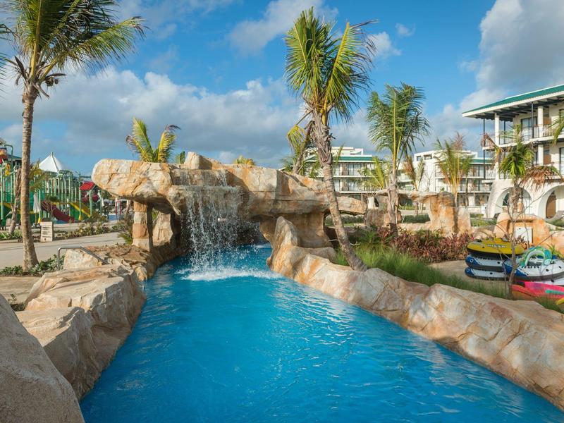 Poolbereich mit Wasserfall, Felsen und Palmen vor einem Hotelgebäude bei sonnigem Wetter.