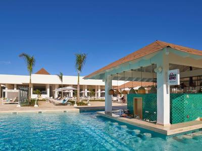 Moderner Pool mit Swim-up-Bar, Palmen und weißem Hotelgebäude unter klarem blauem Himmel.