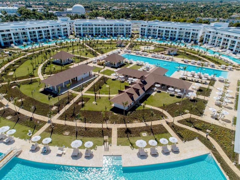 Großes Hotel mit Pool, grünen Rasenflächen, Rundwegen und Sonnenschirmen in weiß und beige.