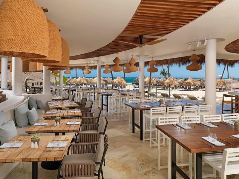 Weites, offenes Restaurant mit Holztischen, Korblampen und Blick auf Pool und Palmenstrand.