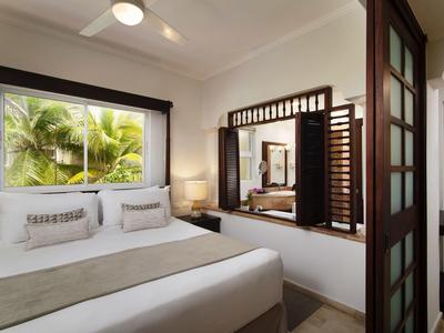 Moderní ložnice s manželskou postelí, okno s výhledem na palmy a velkým zrcadlem na zdi.