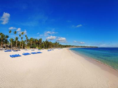 Bílá písečná pláž s modrými lehátky, palmami a čistým modrým mořem pod slunečnou oblohou.