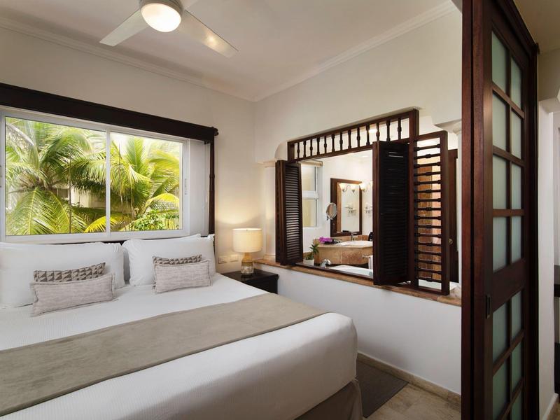 Moderní ložnice s manželskou postelí, okno s výhledem na palmy a velkým zrcadlem na zdi.