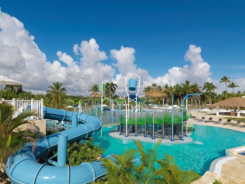 Vodní park s modrými skluzavkami, fontánami a tropickým okolím.