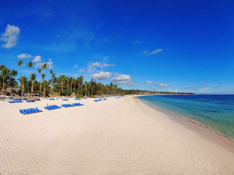 Bílá písečná pláž s modrými lehátky, palmami a čistým modrým mořem pod slunečnou oblohou.