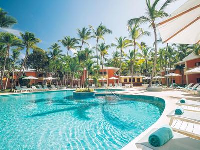 Großer, klarer Pool mit Palmen, Liegestühlen und Sonnenschirmen in einer tropischen Resortanlage.