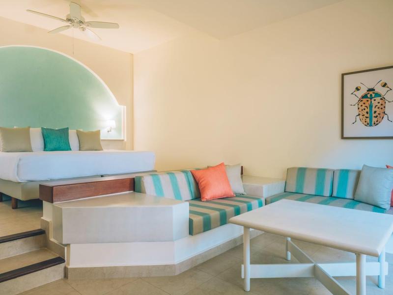 Světly hotelový pokoj s manželskou postelí, barevnými polštáři a posezením s pohovkami a stolem.