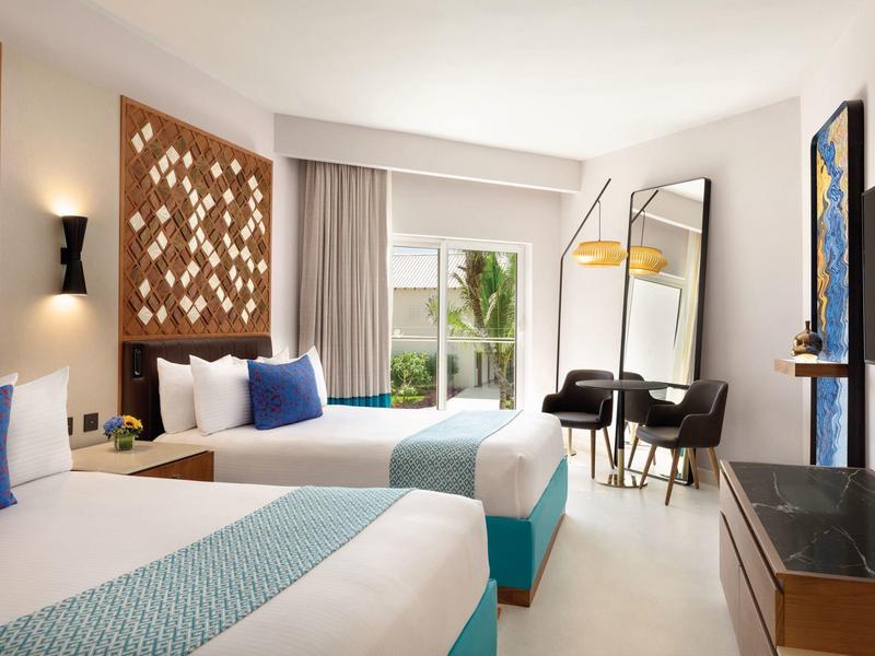 Habitación de hotel luminosa con dos camas, escritorio y gran ventana con vista al jardín.