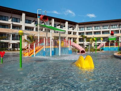 Piscina infantil con toboganes de colores frente a un edificio de hotel de varios pisos bajo cielo azul.