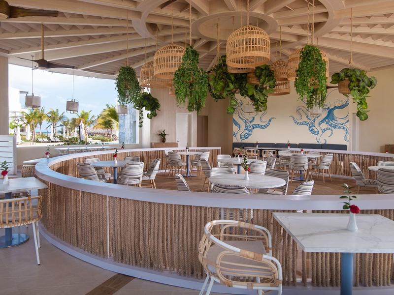 Área abierta de restaurante con sillas de ratán, plantas colgantes y vista a palmeras y al mar.
