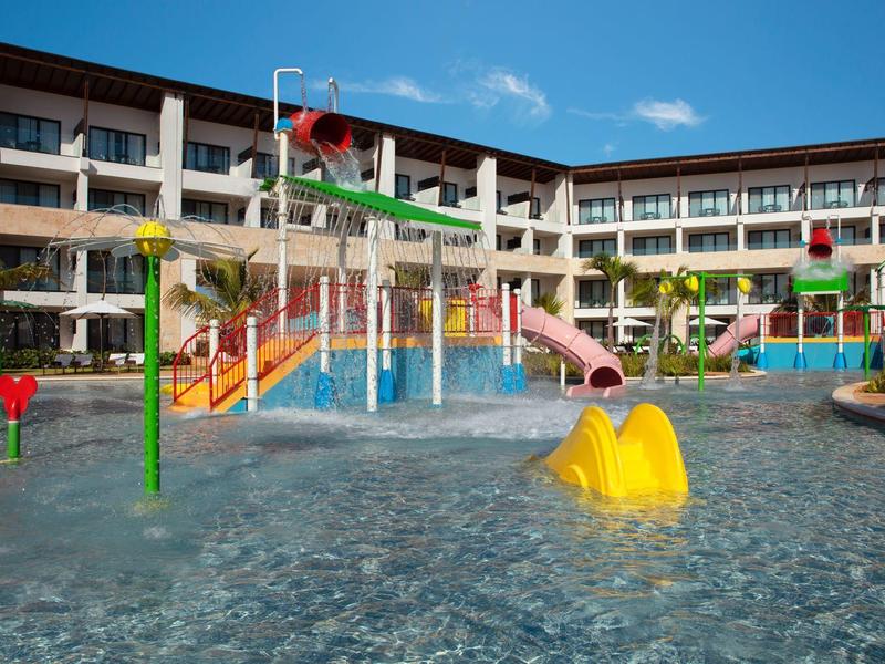 Piscina infantil con toboganes de colores frente a un edificio de hotel de varios pisos bajo cielo azul.