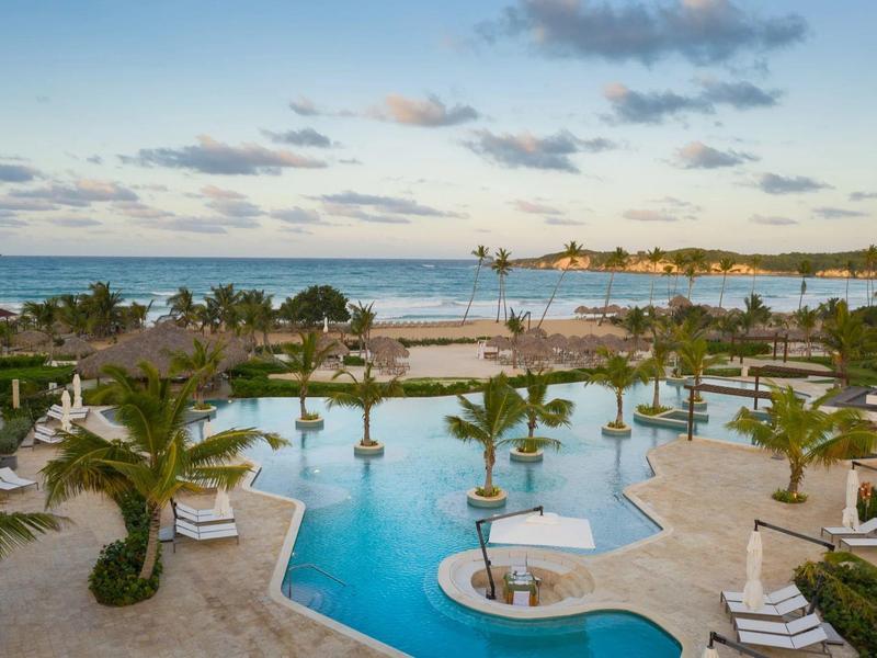 Piscina circular con tumbonas y palmeras cerca de la playa bajo un cielo nublado.