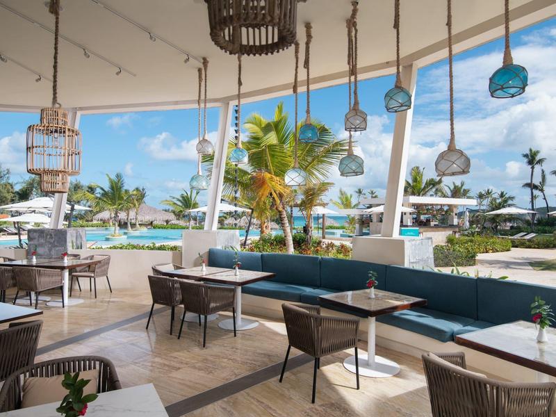 Área de lounge abierta con sillas y mesas con vista a palmeras y piscina en un resort.