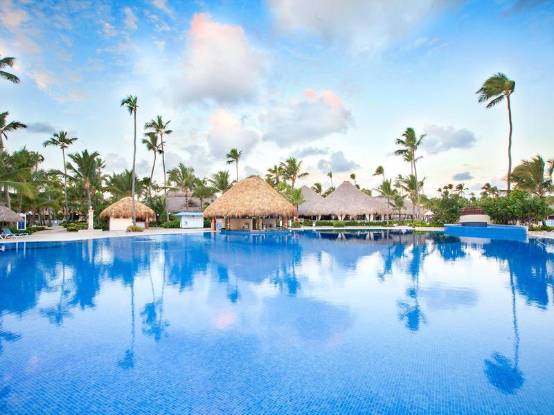Large hotel pool with palm trees and thatched roofs under a blue sky.