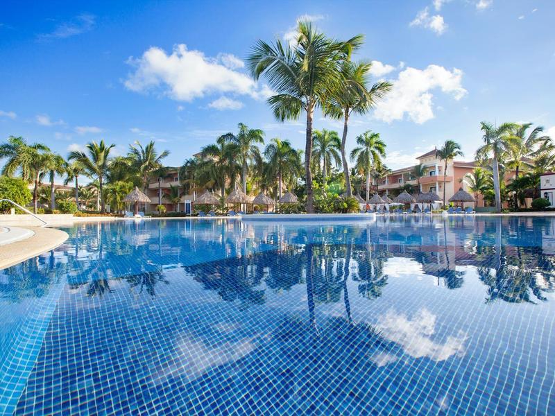 Large pool with reflecting blue water in front of tropical palm trees and holiday homes.