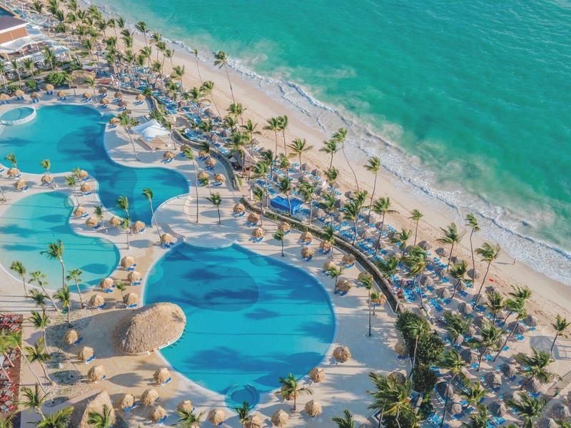 Large pool complex with tropical beach setting and palm trees beside turquoise sea.