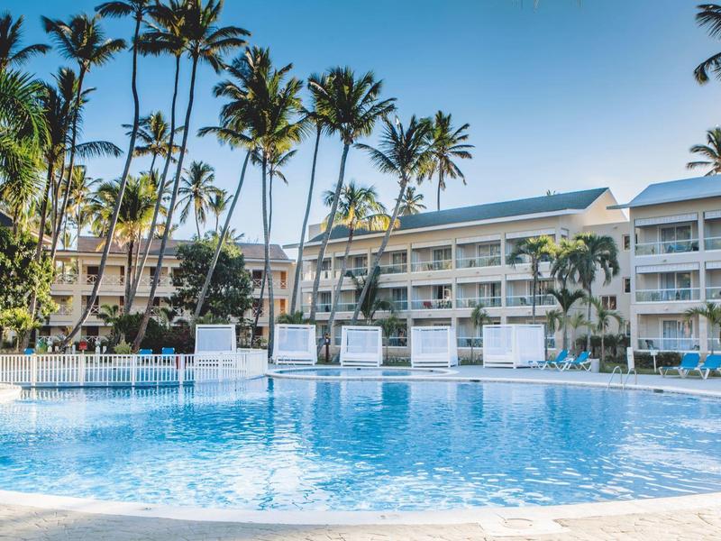Hotel con gran piscina, rodeado de palmeras y edificio blanco de varios pisos al fondo.