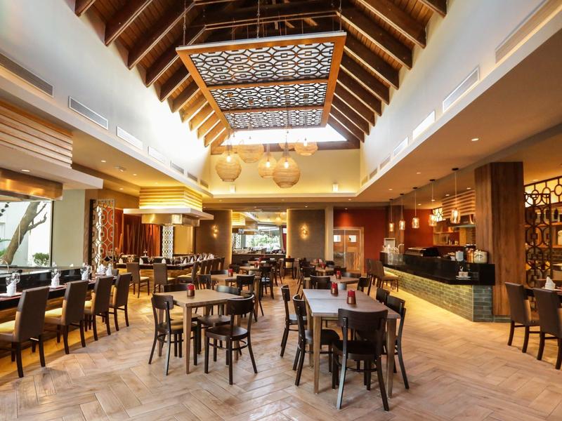 Amplia zona de restaurante con sillas de madera, mesas e interiores claros bajo techo de madera abierto.