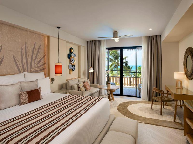 Camera d'hotel luminosa con grande letto, area salotto e balcone con vista sulle palme.