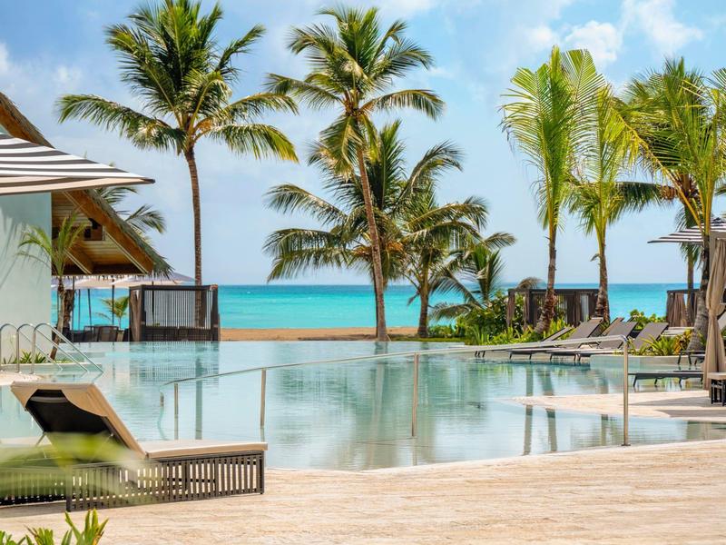 Bazénová zóna s lehátky, palmami a přímým výhledem na moře za jasné oblohy.