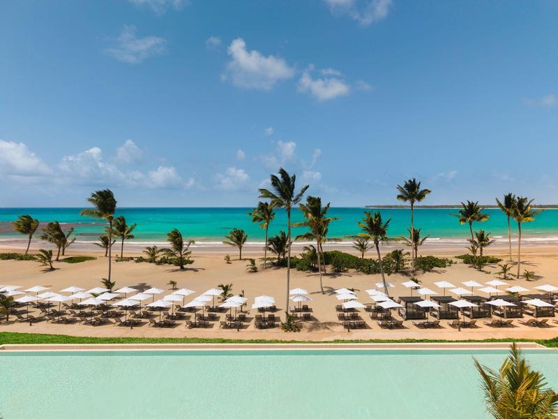 Hotelová pláž s lehátky, slunečníky, palmami a tyrkysovým mořem pod modrou oblohou.