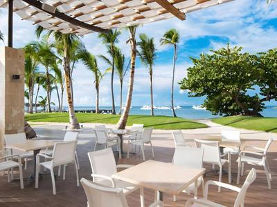 Überdachte Terrasse mit weißen Tischen und Stühlen, Blick auf Palmen und Meer.