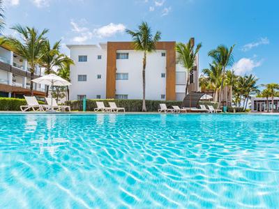 Grande piscina esterna davanti a un hotel moderno con palme e lettini sotto un cielo limpido.