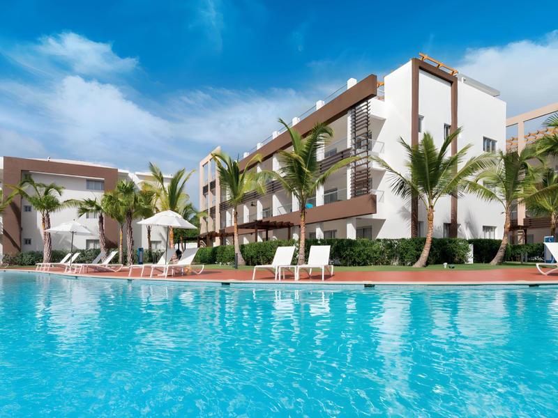 Modern hotelcomplex met groot zwembad, ligstoelen en palmbomen onder een heldere lucht.