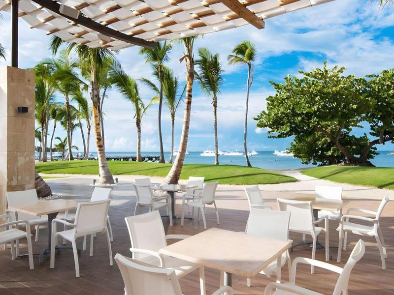 Terras met witte stoelen en tafels, palmbomen en zee op de achtergrond onder blauwe lucht.