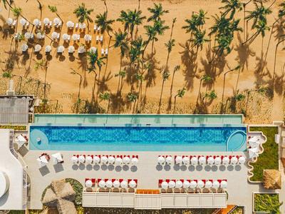 Luftaufnahme eines Hotels mit langem Pool, Liegestühlen, Sonnenschirmen und Palmen am Strand.