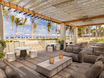 Überdachte Lounge im Freien mit modernen Sitzmöbeln und Blick auf Palmen und Strand.