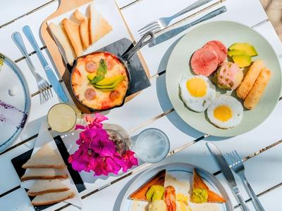 Tavolo con vari piatti per la colazione tra cui uova, toast e bevande, illuminato da luce intensa.