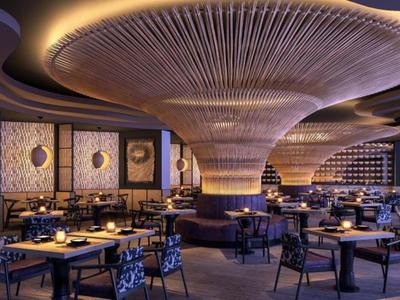 Elegancka restauracja hotelowa z nowoczesnym designem i ciepłą atmosferą, wieloma stołami i efektownym oświetleniem sufitowym.