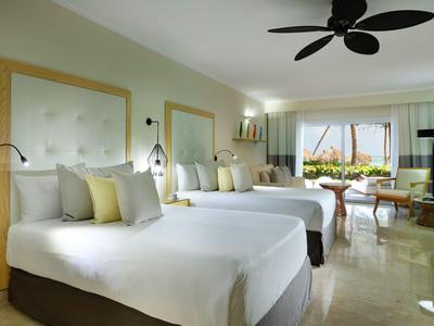 Helles Hotelzimmer mit zwei Doppelbetten, Ventilator an der Decke und Blick auf den Garten.